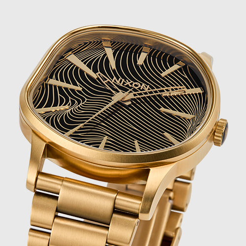 A close-up shot of a gold Nixon watch with a black face featuring intricate gold wavy lines and a gold band.