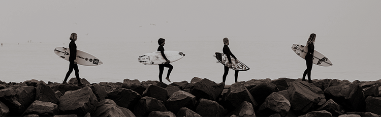 Four surfboarders walk along the ocean while holding their surfboards.